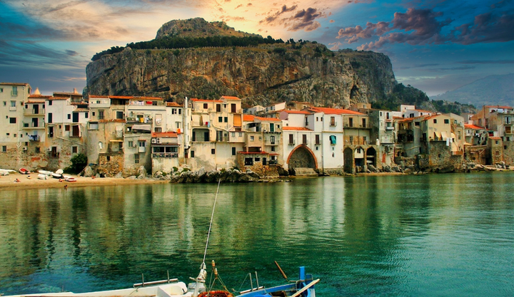 cenic coastal view of Cefalù, Sicily, with historic medieval buildings along the waterfront, calm turquoise sea, and towering rocky mountain backdrop under a dramatic sunset sky.