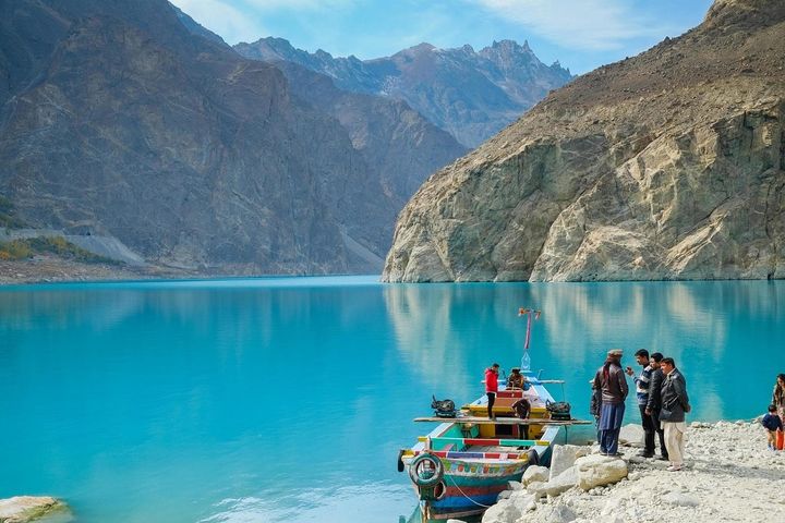 Men gathered near a boat on the Hunza river in Pakistan, which is a bright blue river surrounded by tall mountain peaks.