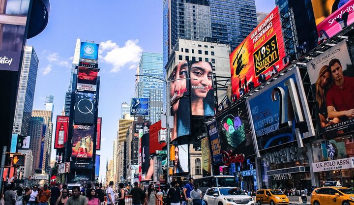 Tourist walking around Time Square in New York City with bright colored advertisements on the sides of buildings and cars passing by.