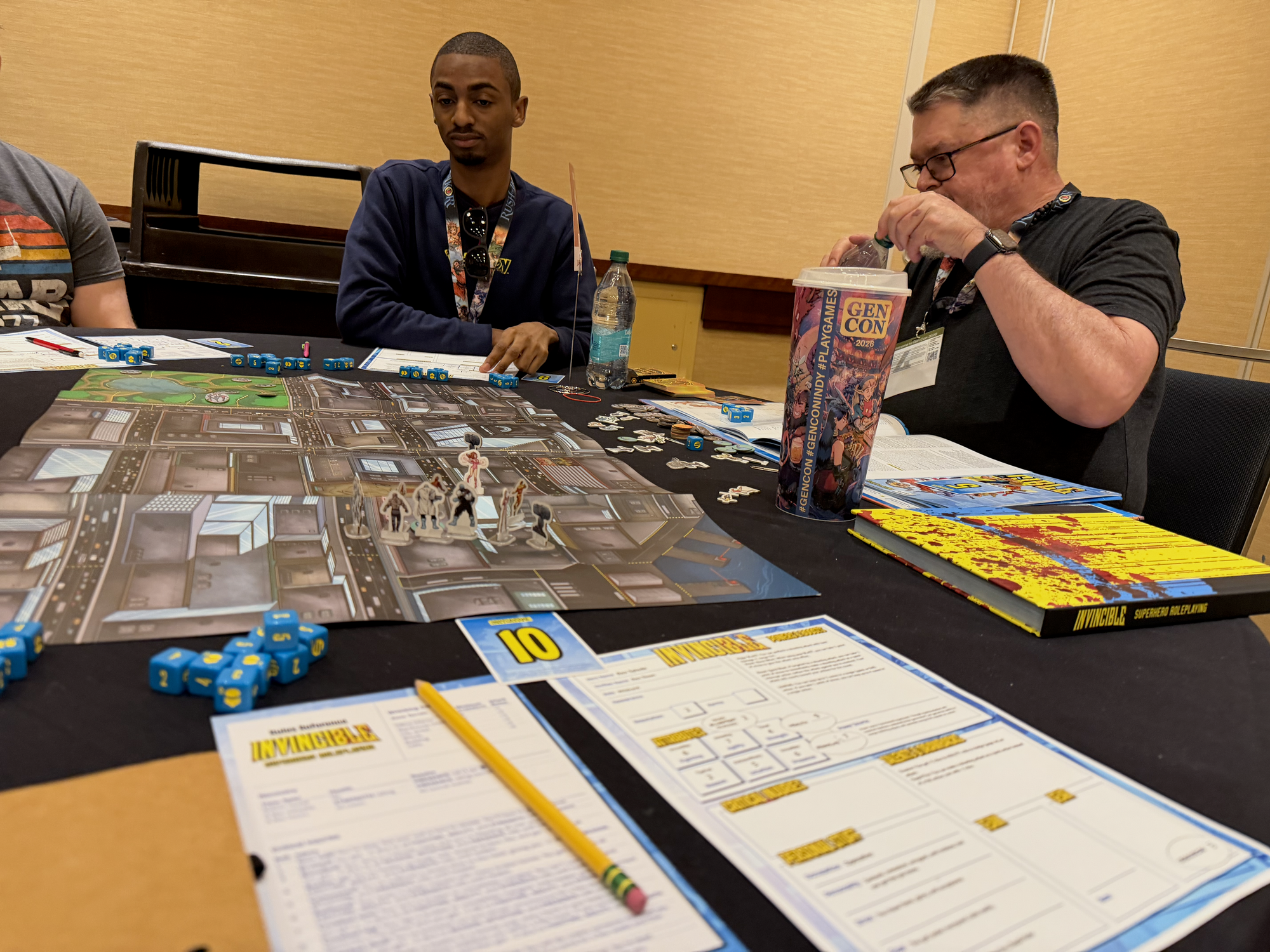 Two guys at a table, with a city maps, card standees, and the Invisible core book on the table. Cool blue and yellow dice.