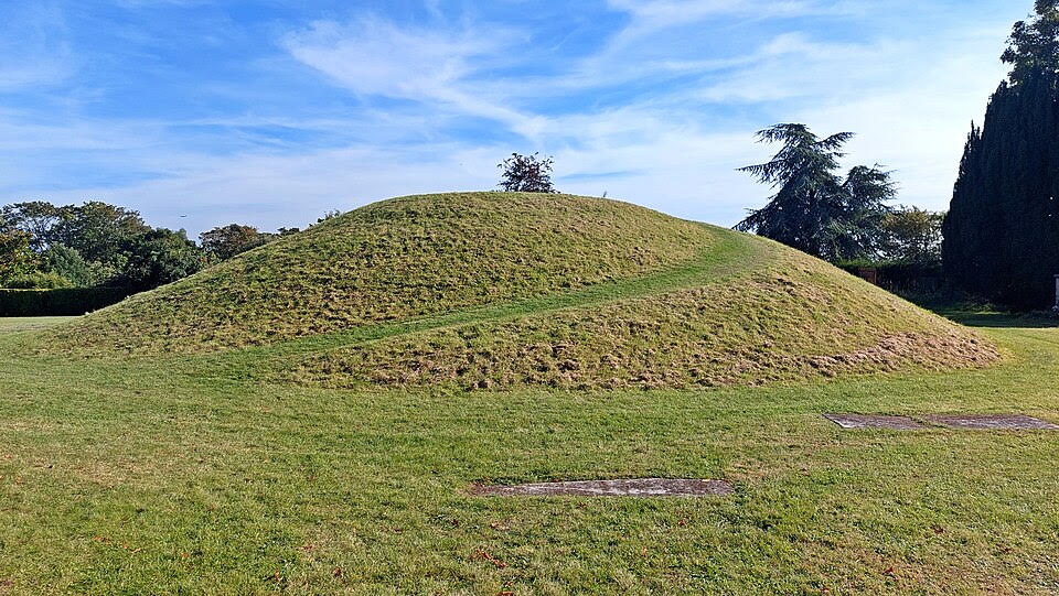 A green barrow mound rises from a lightly wooded area under a blue sky and fleecy clouds. A path, a deeper green, spirals up to the top.