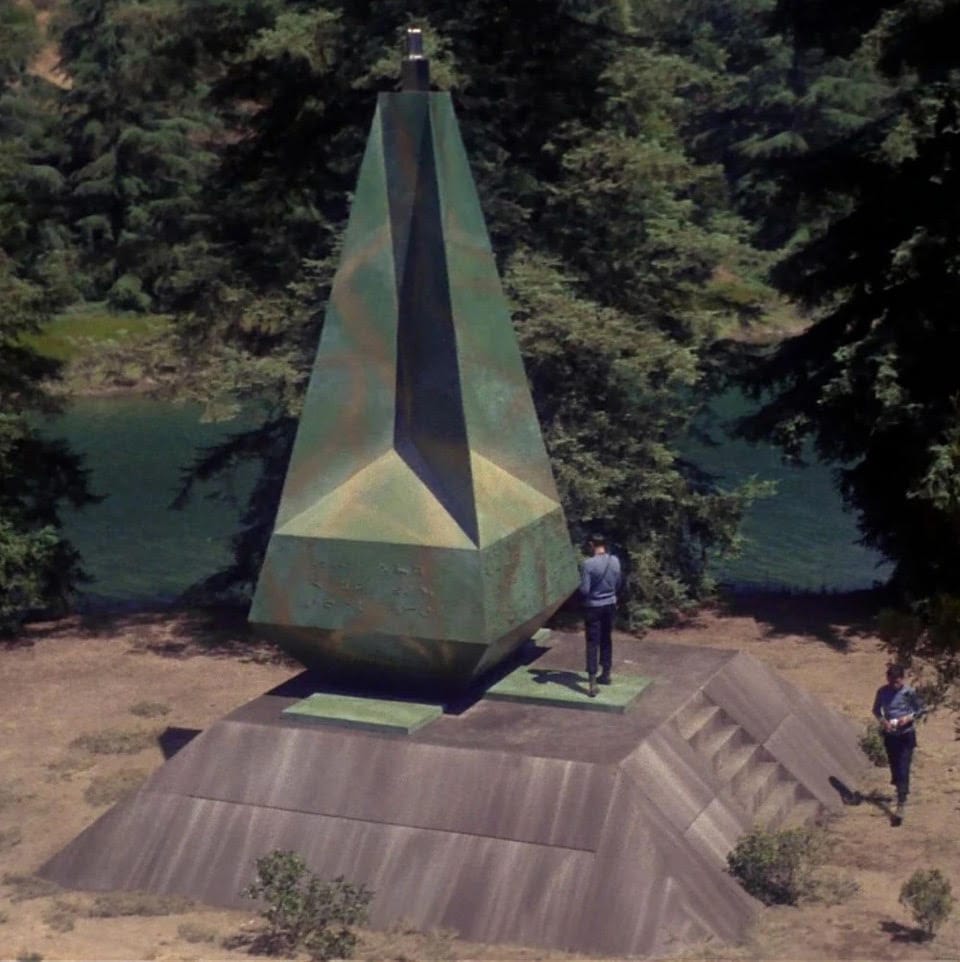 Spock and McCoy, in a woodland outdoor setting beside a river, are investigating an alien structure. There is a huge grey platform supporting a very tall, metallic looking object that looks somewhat like a tapered four-sided pyramid, but with many indentations and covered in alien writing along the widest part. The object is a pale green color but with golden swirls amid the green.