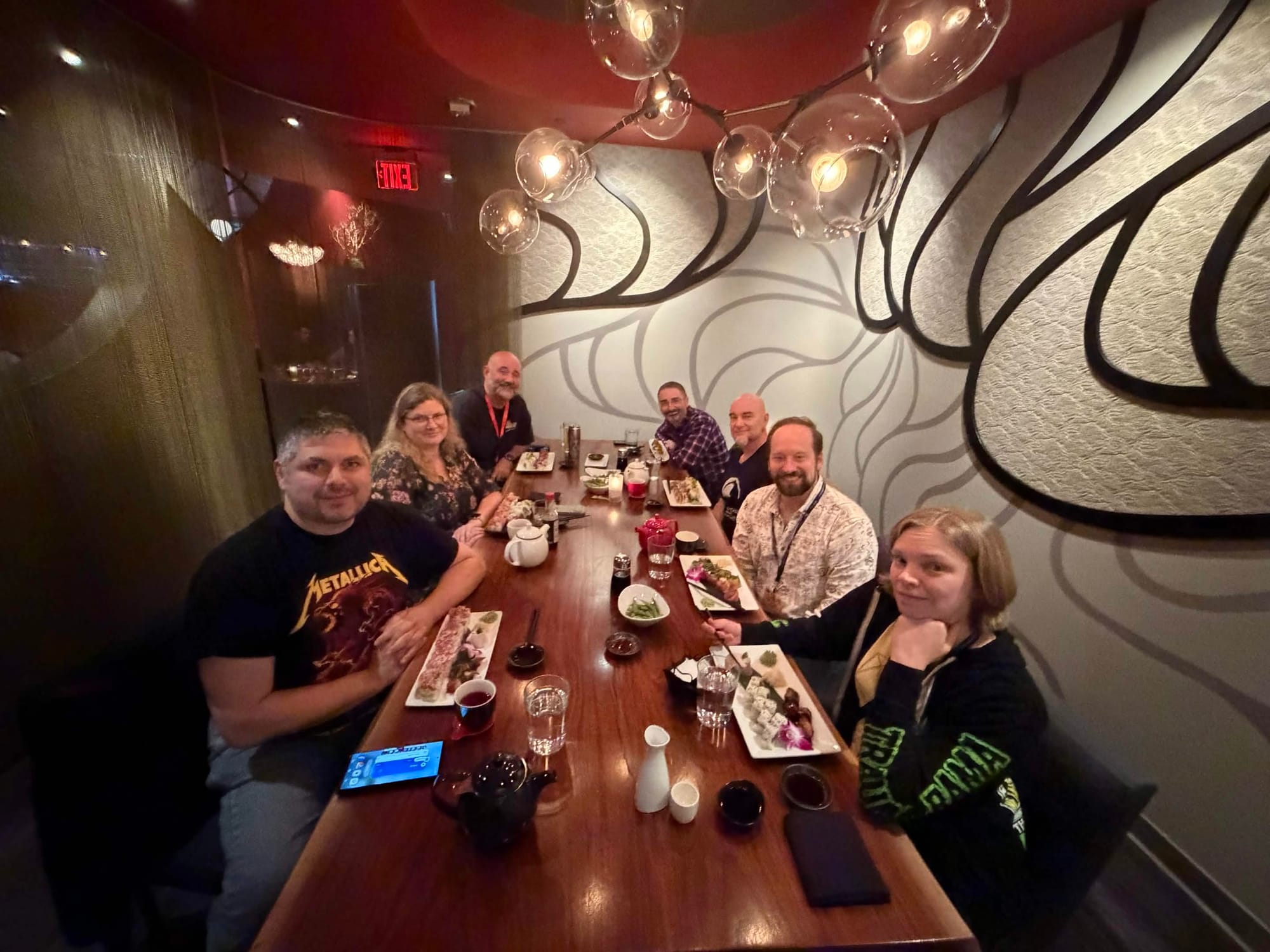 Seven folks seated in a private room on a polished red wood table eating sushi