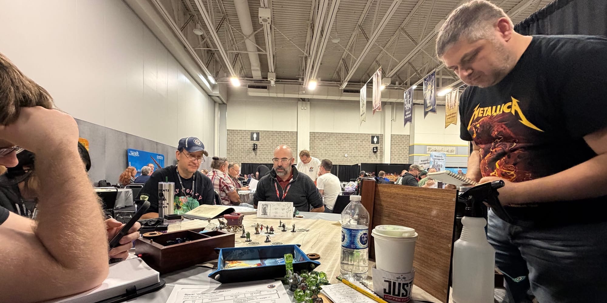 Andrew Bishkinskyi standing on the right over a wooden GM's screen. Three other players visible, with dice and minis on a table.