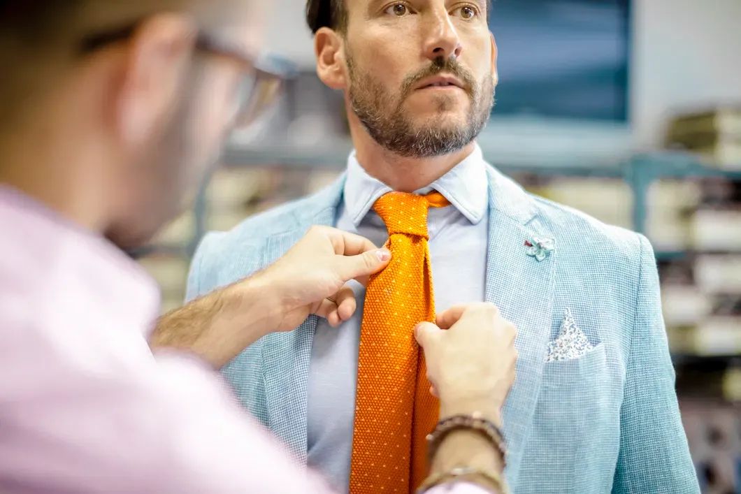 Tailor adjusting an orange tie on a man wearing a light blue linen jacket and pocket square – Derby Day style and linen jacket fit for the Kentucky Derby.