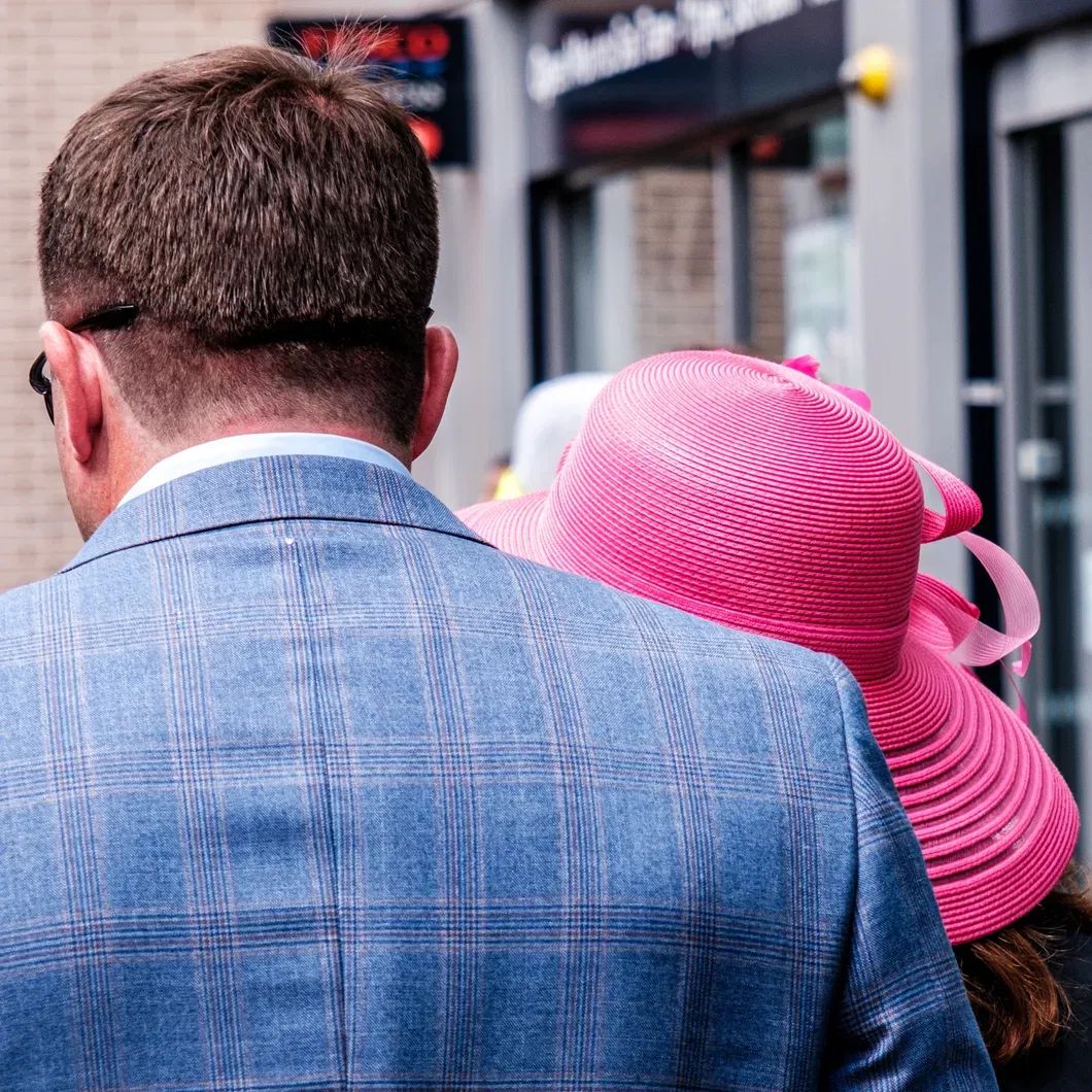 Back view of a man in a blue checked suit jacket and a woman in a bright pink hat, stylishly dressed for a British summer racing event.