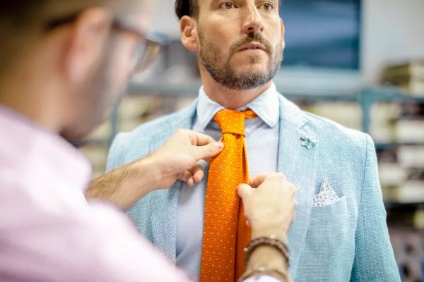 Tailor adjusting an orange tie on a man wearing a light blue linen jacket and pocket square – Derby Day style and linen jacket fit for the Kentucky Derby.