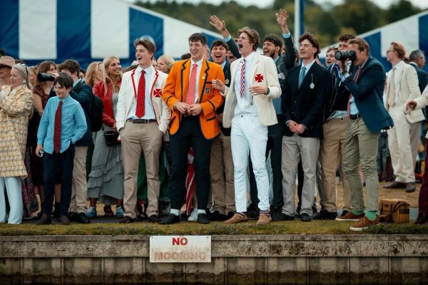 A lively group of young men in colourful rowing blazers, ties, and summer suits cheering on the riverbank at Henley Royal Regatta, with crowds and blue-and-white marquees behind them.