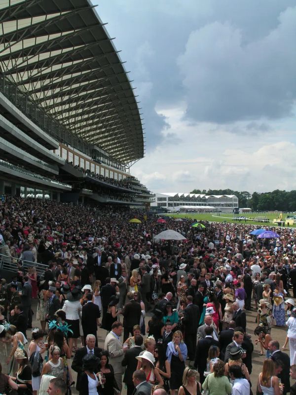 Large crowd in formal attire at Royal Ascot racecourse, with grandstand and dramatic sky during the British social season.
