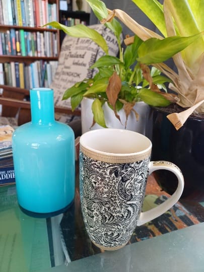 A cup, patterned with a black and white botanical design, standing on a glass coffee table with a blue vase and two plants grouped behind it.