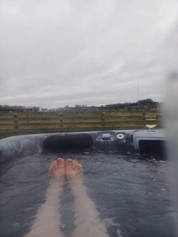 A view from someone holding a camera in a spa pool, showing her bare legs floating in the water. There's a glass of wine on the rim, and beyond is a wide horizoned landscape of farmland and grey sky.
