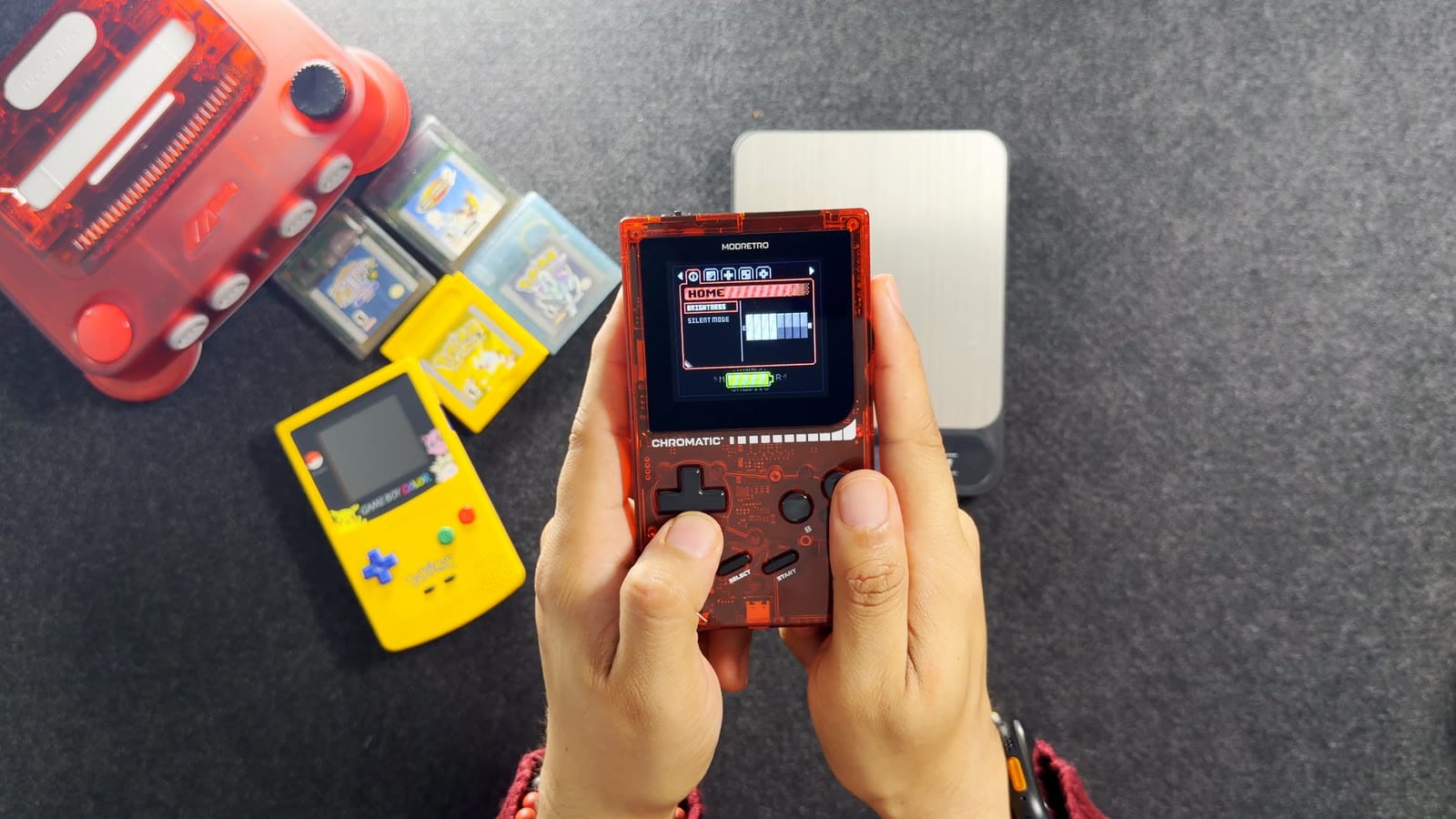 The Chromatic held in two hands with its settings menu on screen, a Game Boy Color on the mat beside it