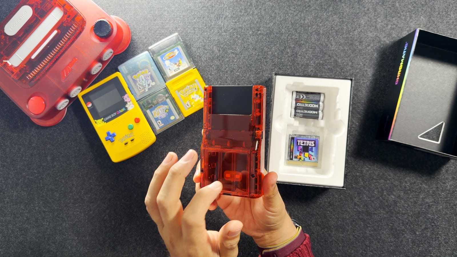 Lifting the transparent red Chromatic out of its box, with three ModRetro AA batteries and a transparent Tetris cartridge in the tray