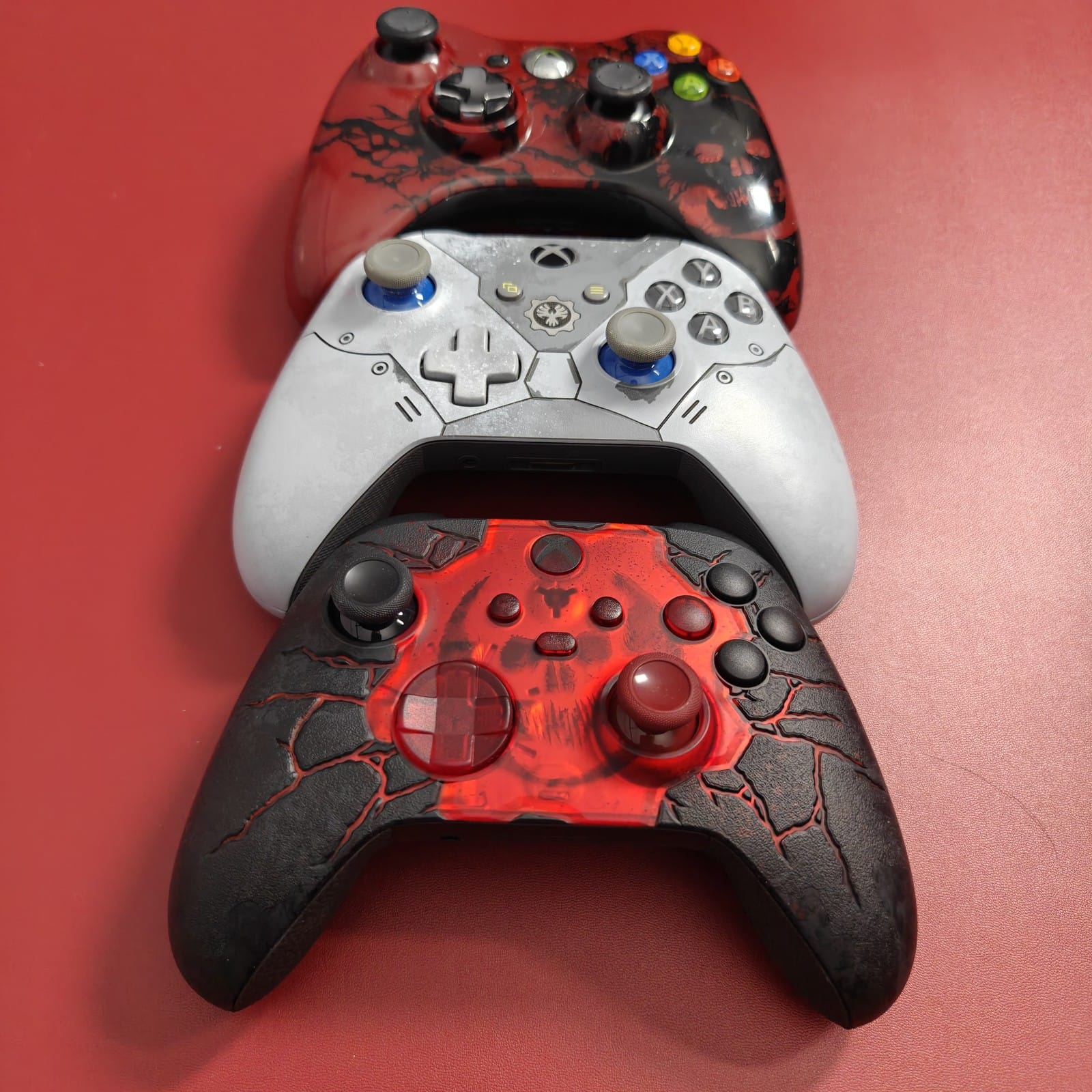 Three Gears of War Xbox controllers stacked: the red and black Gears of War 2 controller, the grey Gears 5 controller and the E-Day controller