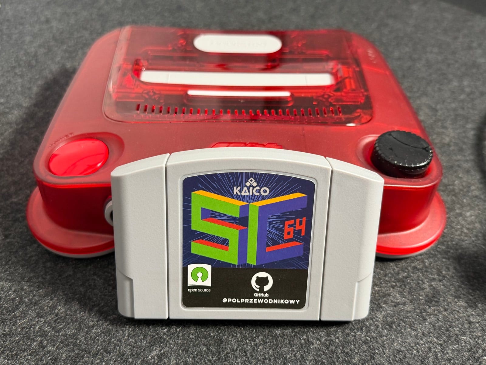A grey SummerCart64 cartridge with a Kaico label standing in front of a translucent red ModRetro M64