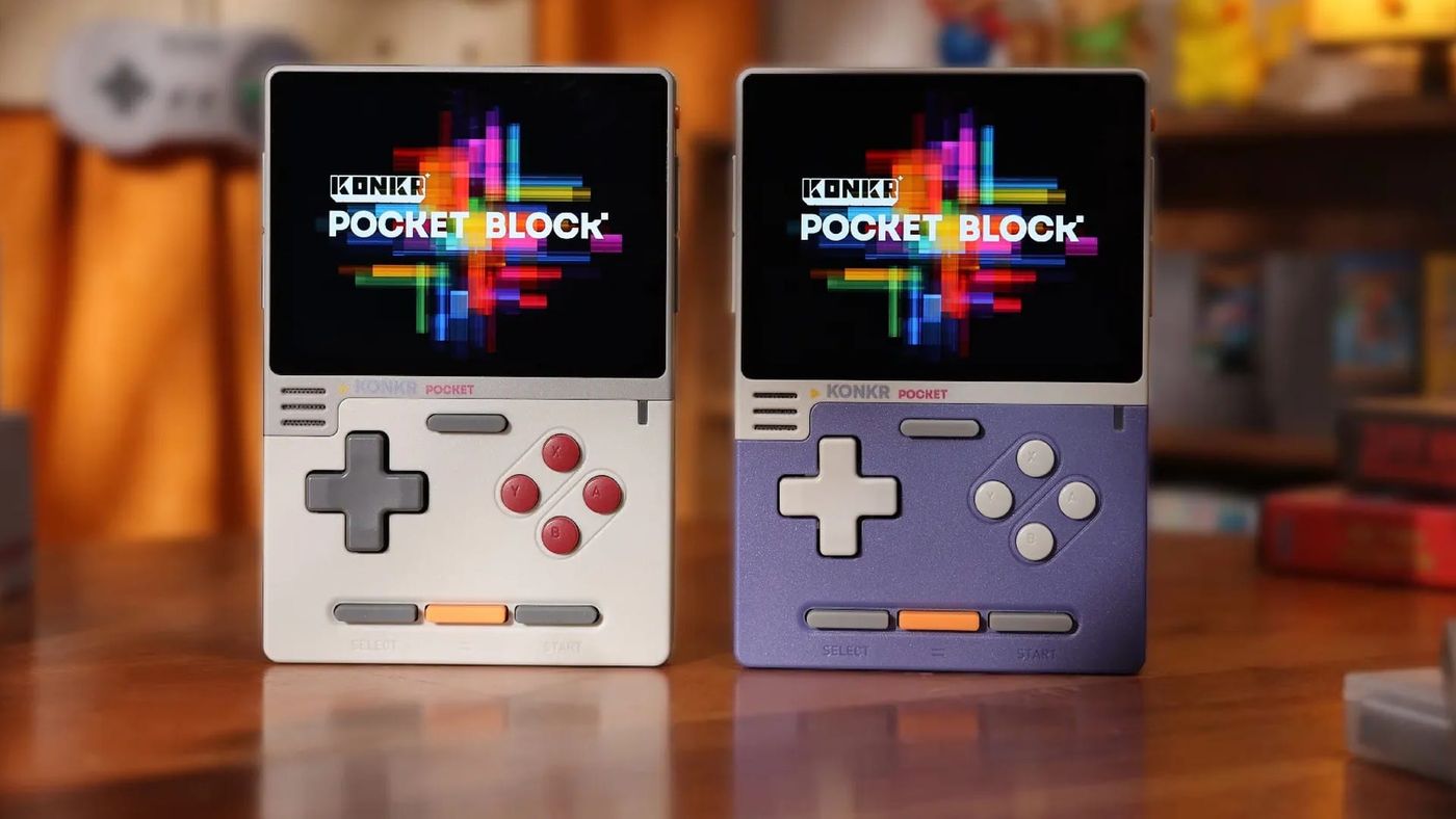 Two KONKR Pocket Block handhelds, grey and purple, standing on a wooden table in front of retro games