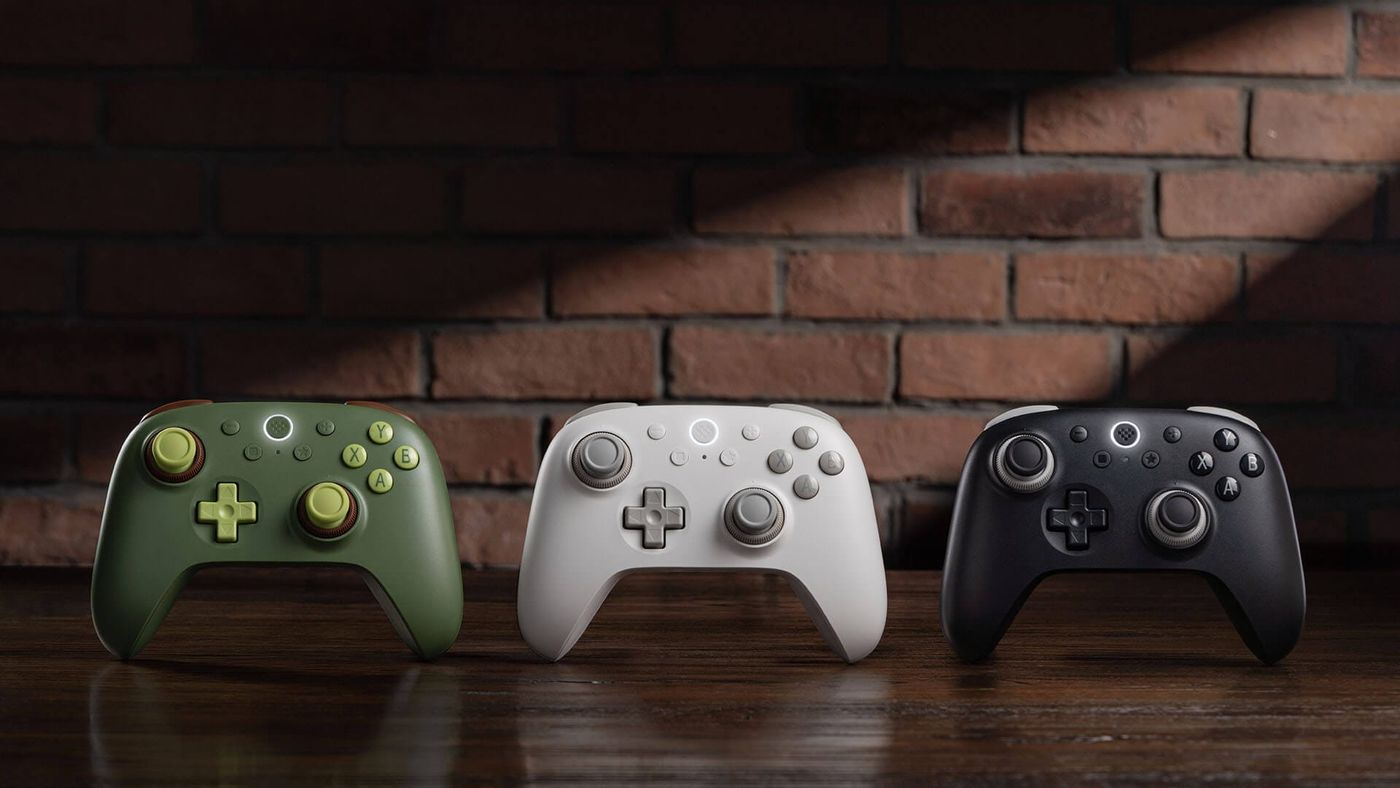 The 8BitDo Ultimate 3C+ in Avocado Green, Frost White and Stealth Black side by side on a wooden table