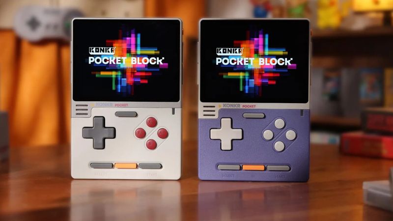 Two KONKR Pocket Block handhelds, grey and purple, standing on a wooden table in front of retro games