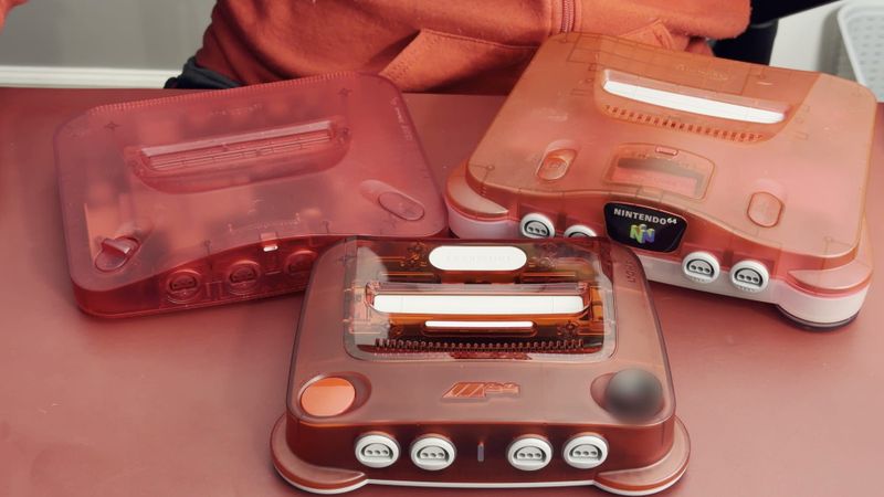 A transparent red ModRetro M64 in front of a transparent red Analogue 3D and an original red Nintendo 64