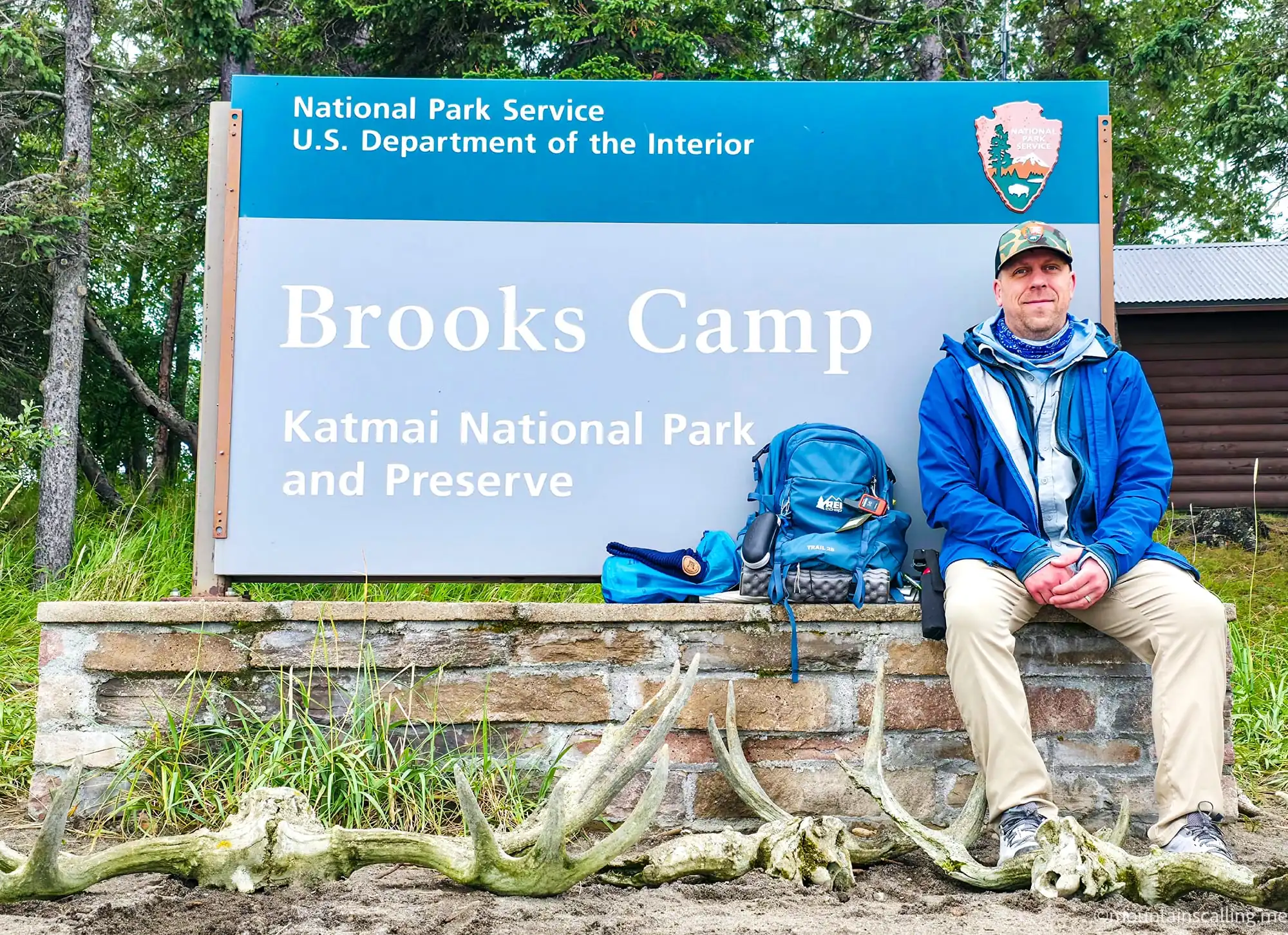 Eric Kufrin sitting at Brooks Camp sign in Katmai National Park with backpack and caribou antlers on ground
