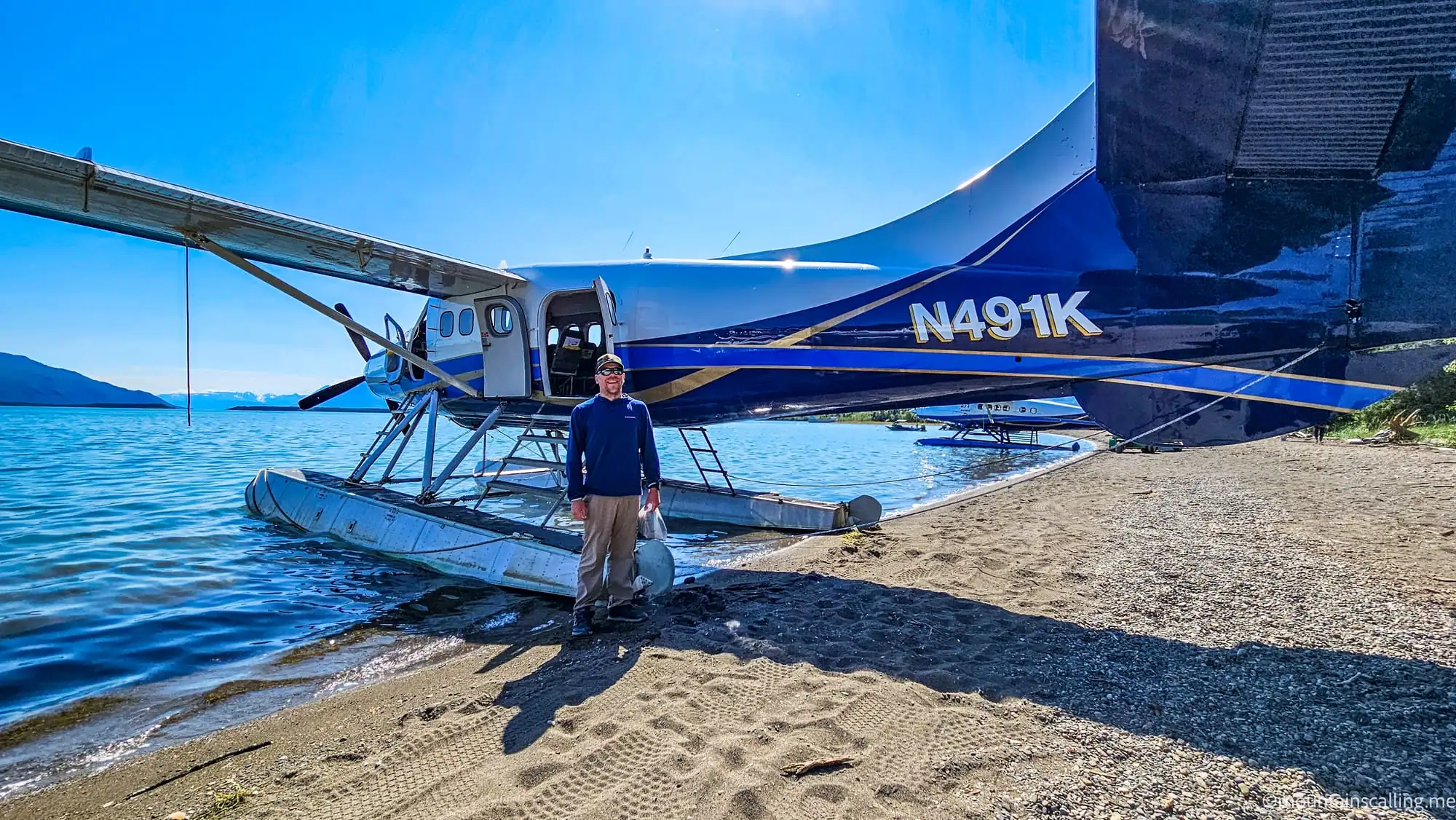 Floatplane on beach with pilot standing beside aircraft, mountains and water in background