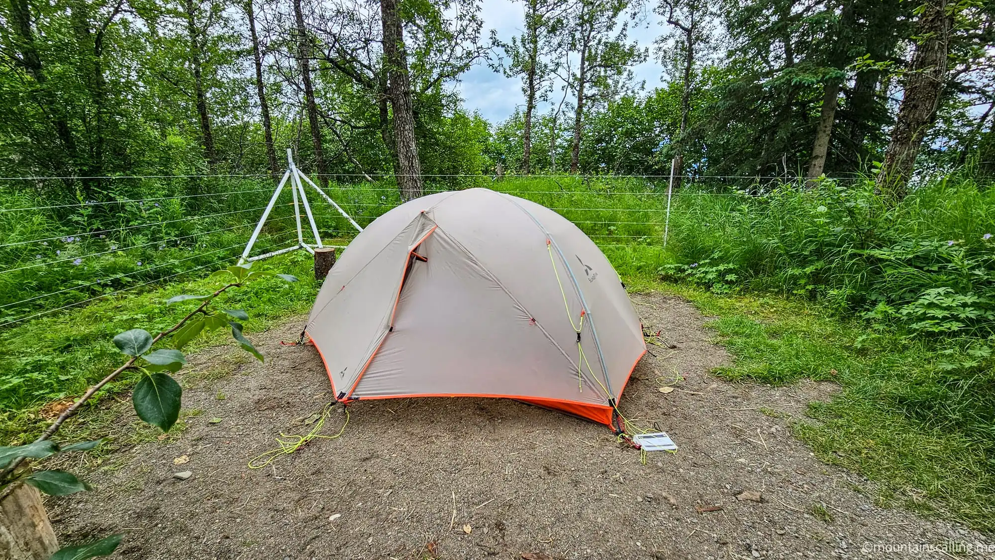 Gray camping tent set up on gravel pad surrounded by electric fence wires in wooded campground