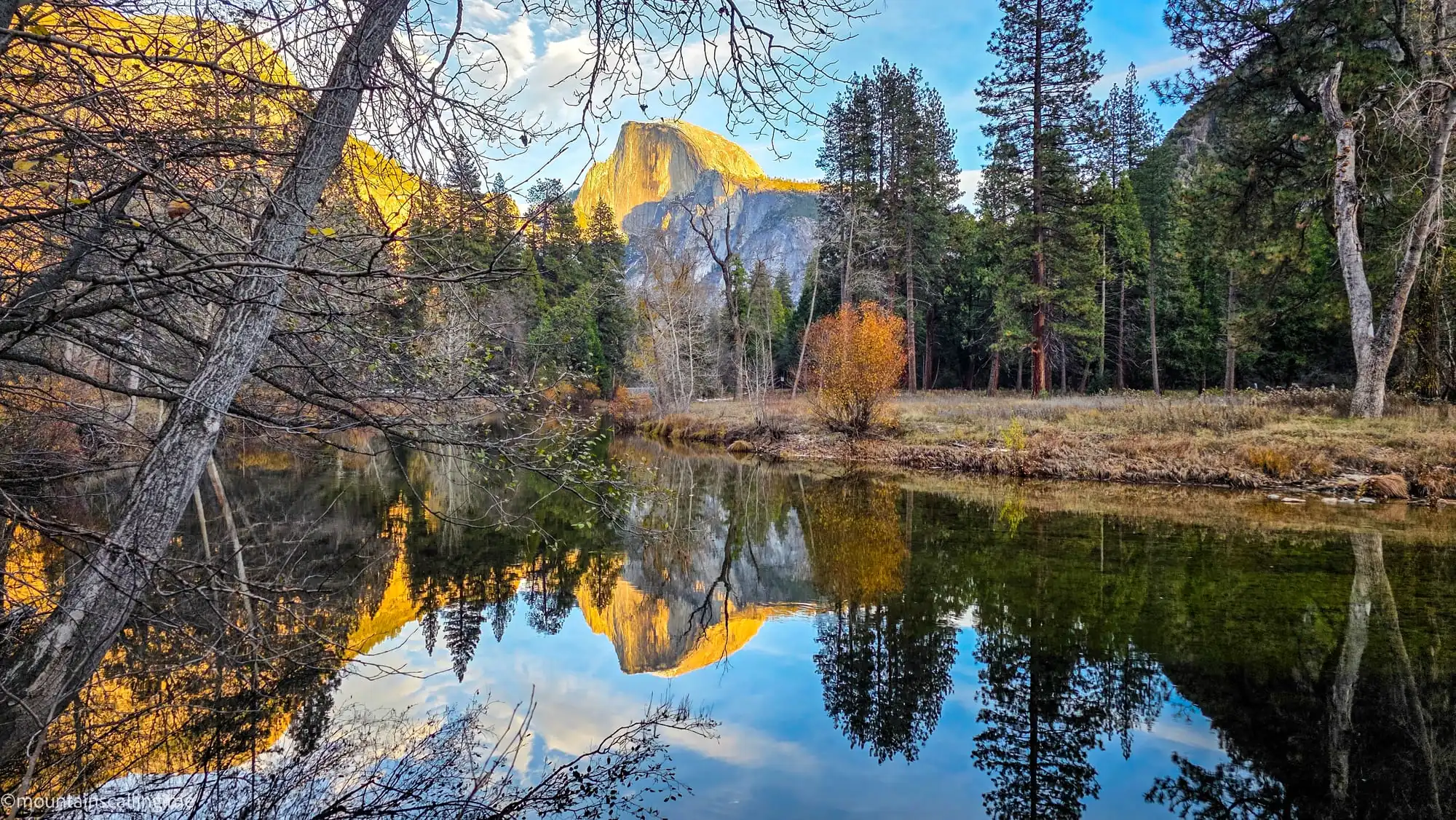 Half Dome's granite face reflected in calm waters of Merced River during golden hour in Yosemite Valley with autumn trees