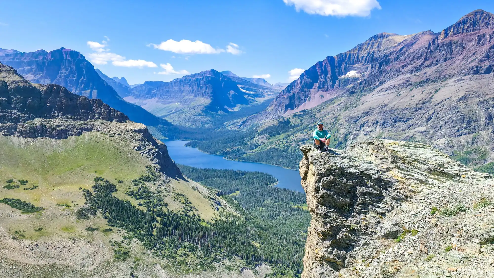 Hiker sitting on rocky cliff overlooking mountain valley and lake in Glacier National Park Montana