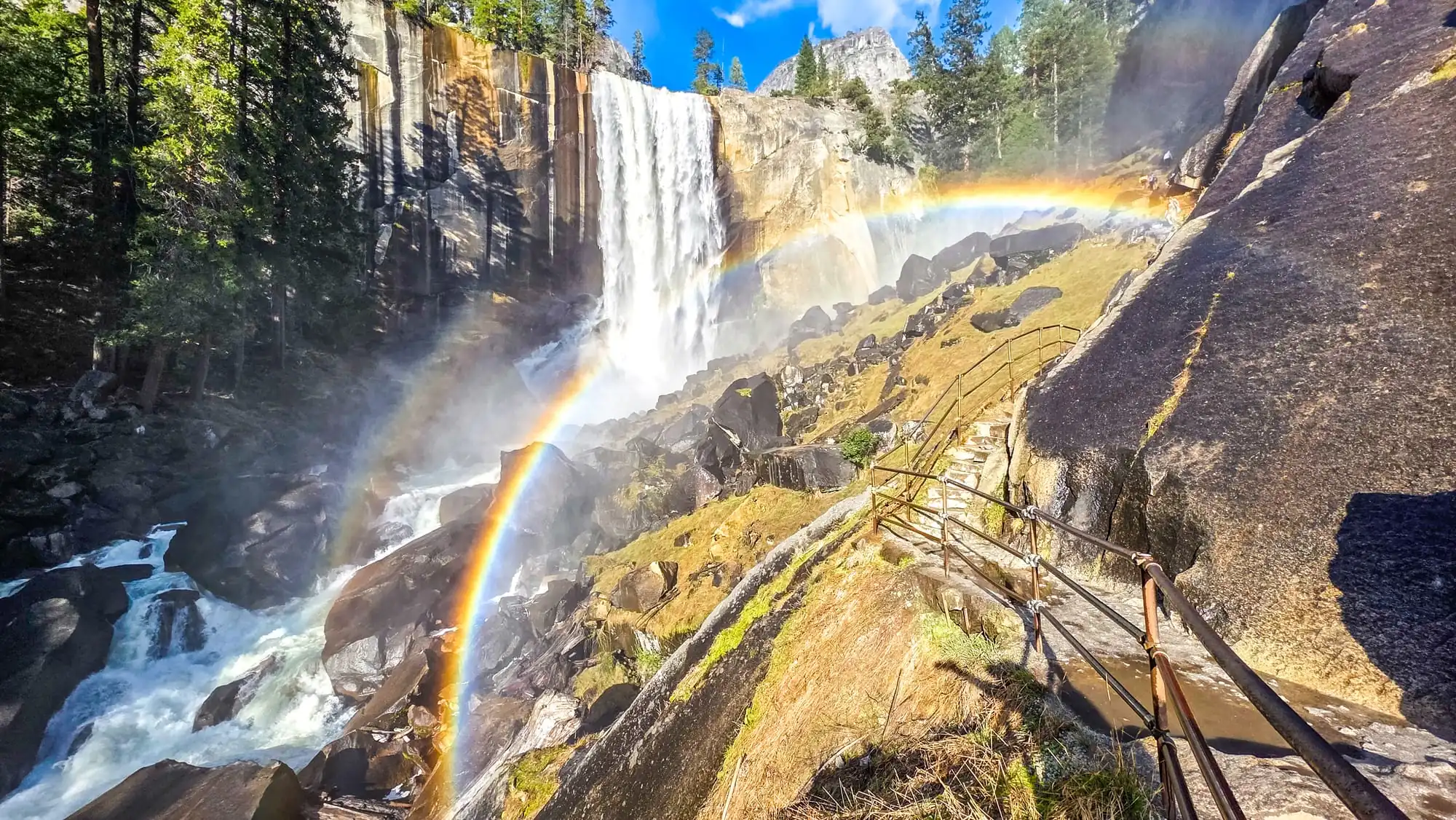 Dramatic waterfall cascades down granite cliffs with double rainbow arcing through mist and spray, wooden trail leading to viewpoint