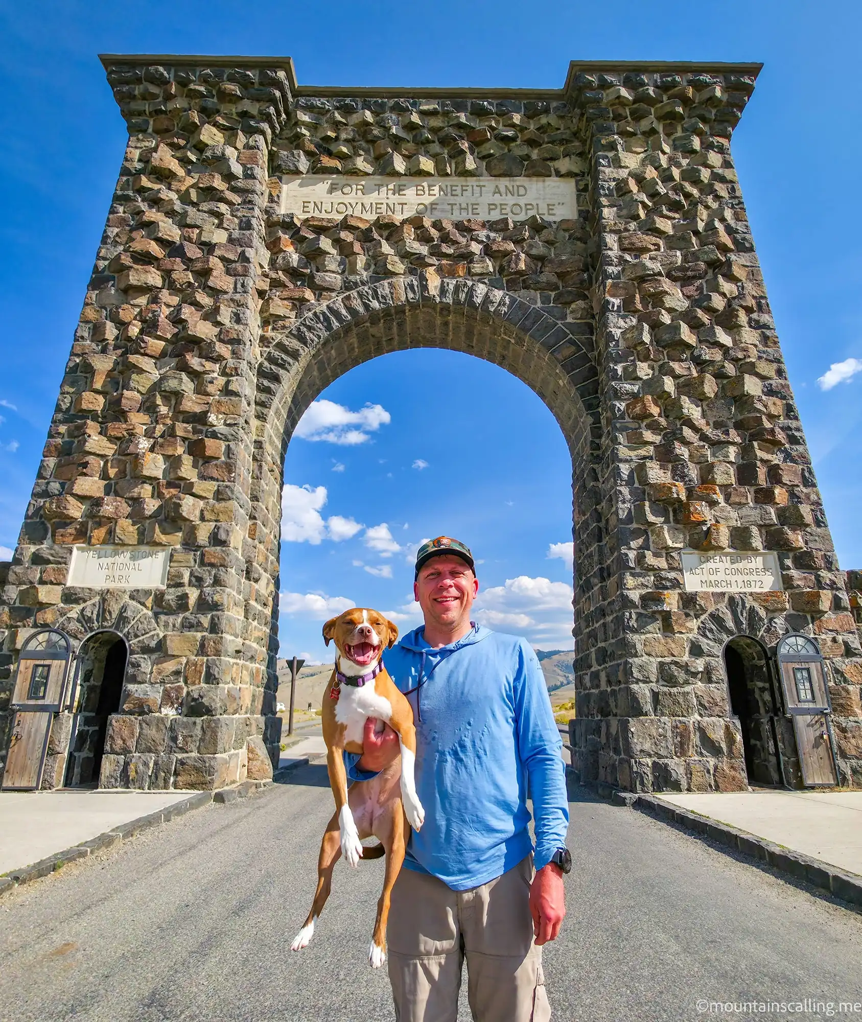 Eric Kufrin and his dog Kali standing beneath the Roosevelt Arch stone entrance to Yellowstone National Park on a clear blue sky day