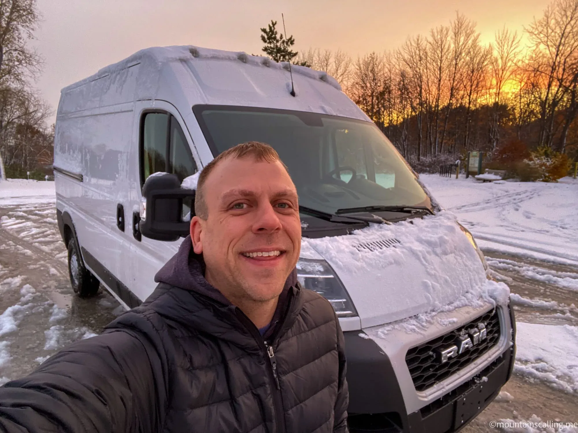 Eric Kufrin taking a selfie in front of his white Ram ProMaster van in snowy winter conditions at sunset