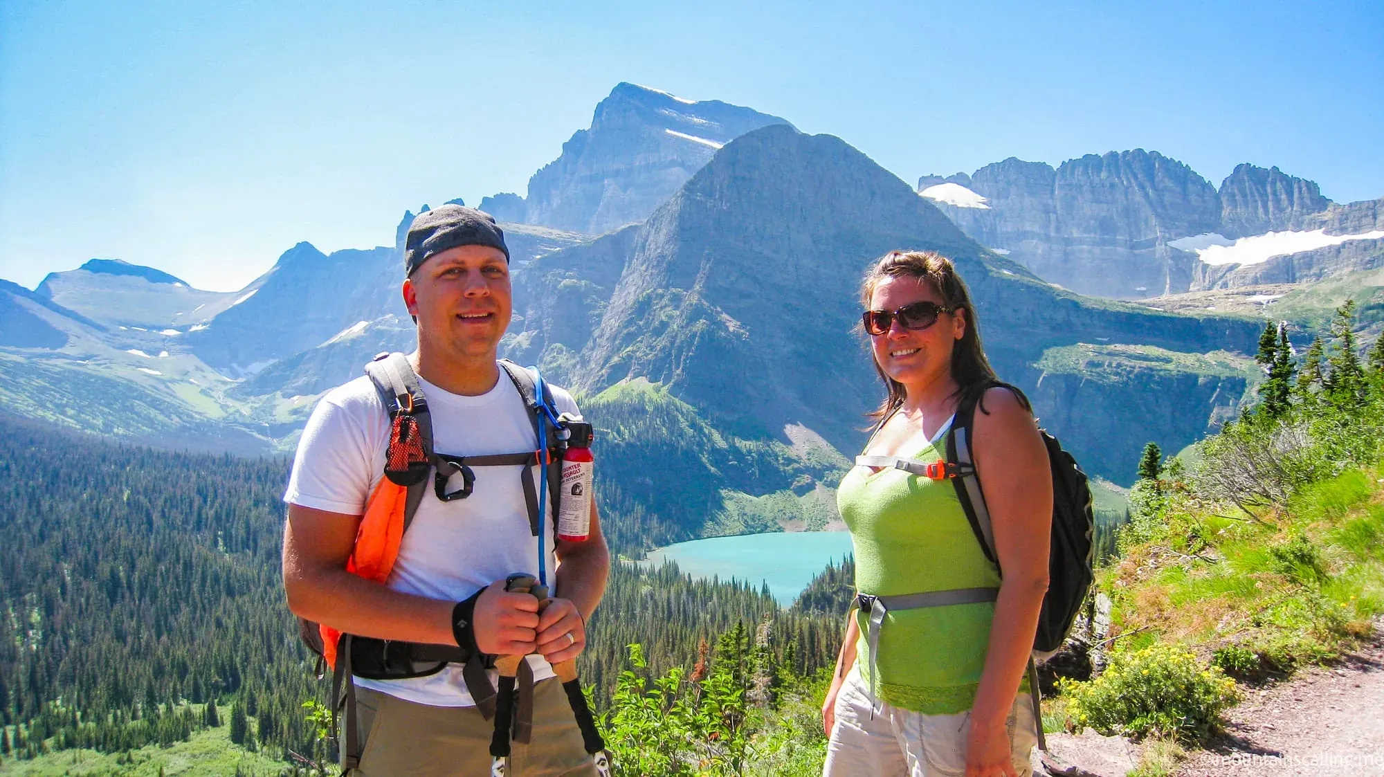 Eric Kufrin and Valerie with backpacks and trekking poles posing on Grinnell Glacier Trail with mountain lake and glaciated peaks behind them in Glacier National Park