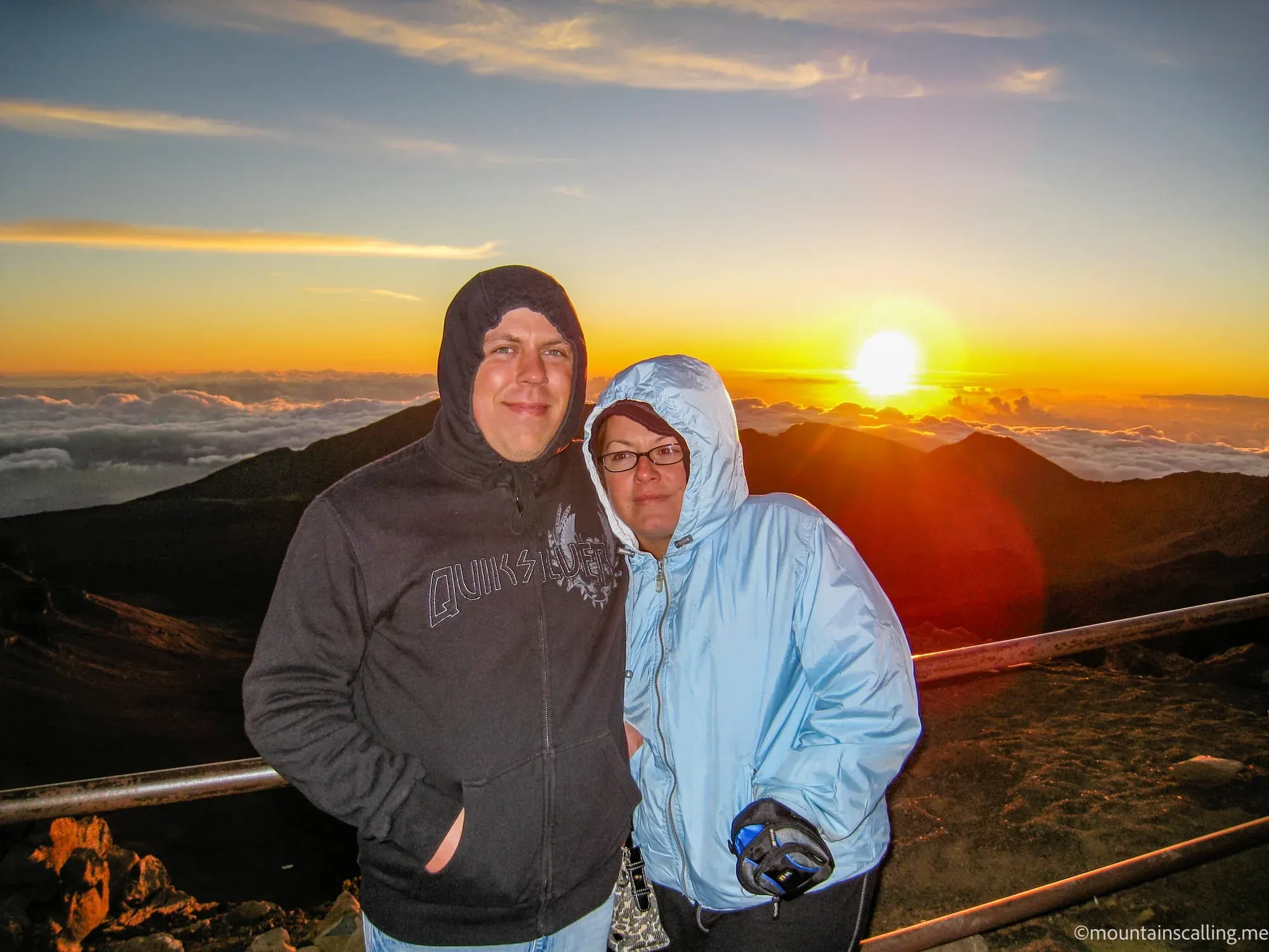 Eric Kufrin and Valerie in hooded jackets watching sunrise from Haleakalā summit with volcanic landscape and clouds below