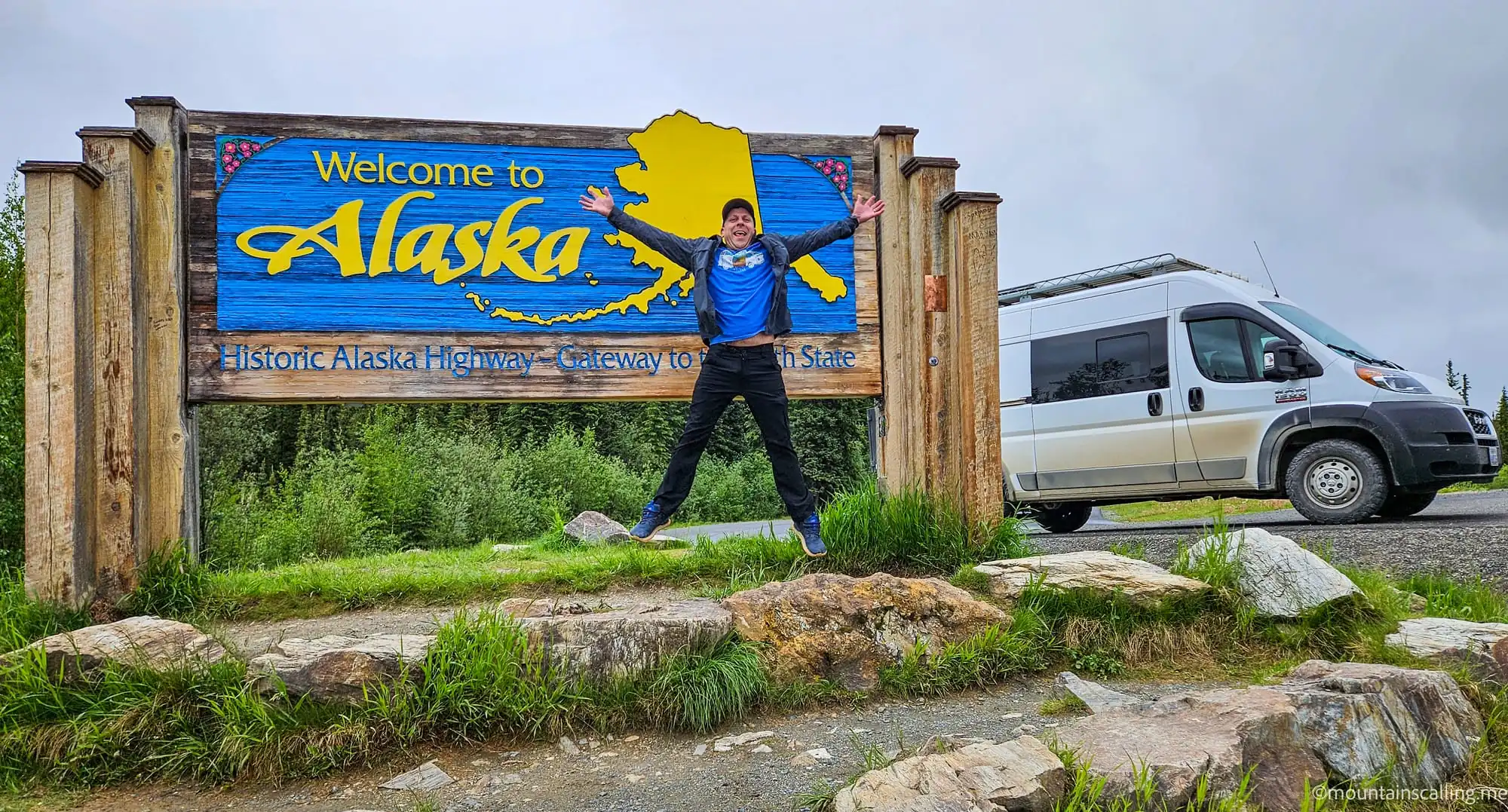 Eric Kufrin jumping excitedly in front of Welcome to Alaska highway sign with RV parked nearby