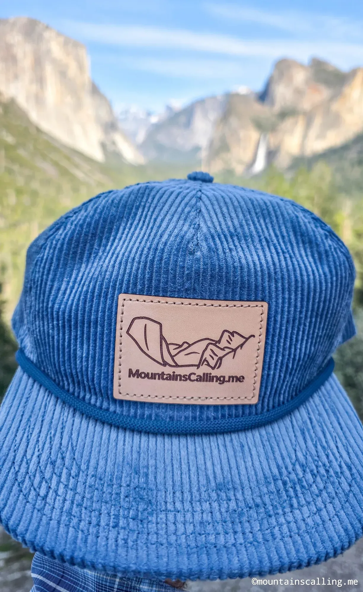 Blue corduroy trucker hat with MountainsCalling.me leather patch against blurred Yosemite Valley granite cliffs