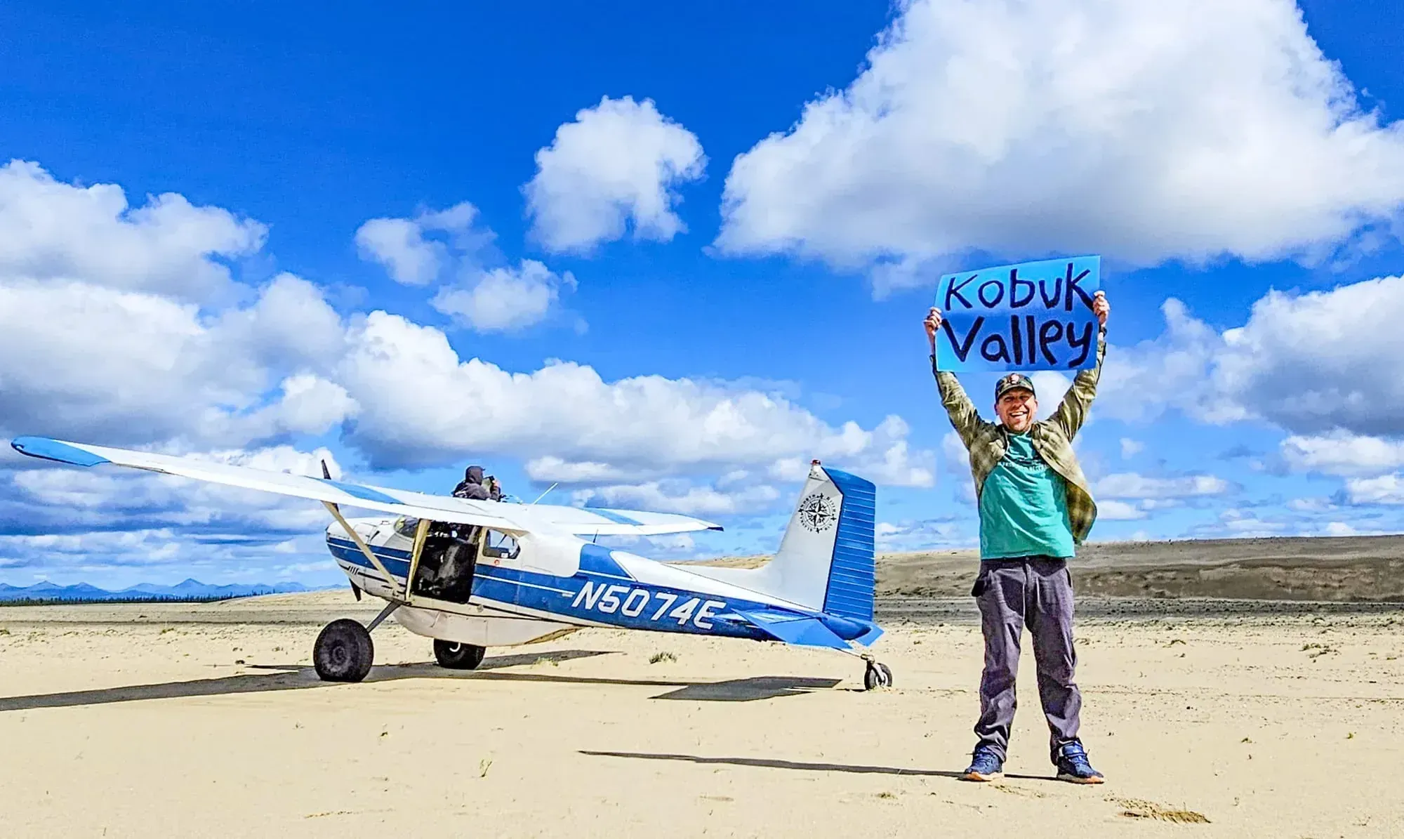 Eric Kufrin holding Kobuk Valley sign next to bush plane on remote Alaska airstrip with dramatic sky and wilderness landscape