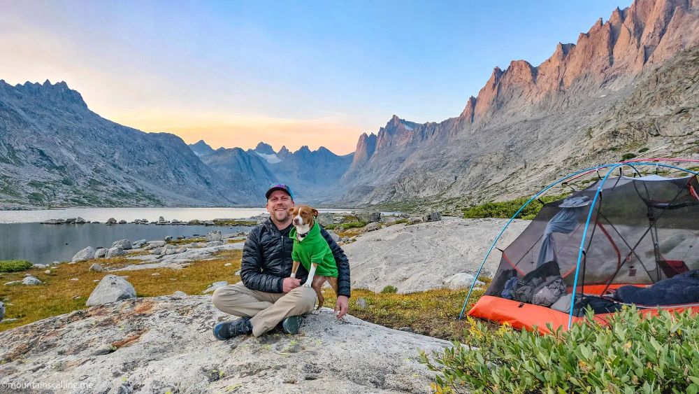 Eric Kufrin sitting with his dog Kali beside camping tent at mountain lake during sunset, rocky peaks in background