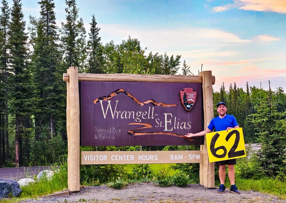 Eric Kufrin holding yellow 62 sign at Wrangell-St Elias National Park entrance, standing next to wooden park sign with visitor center hours