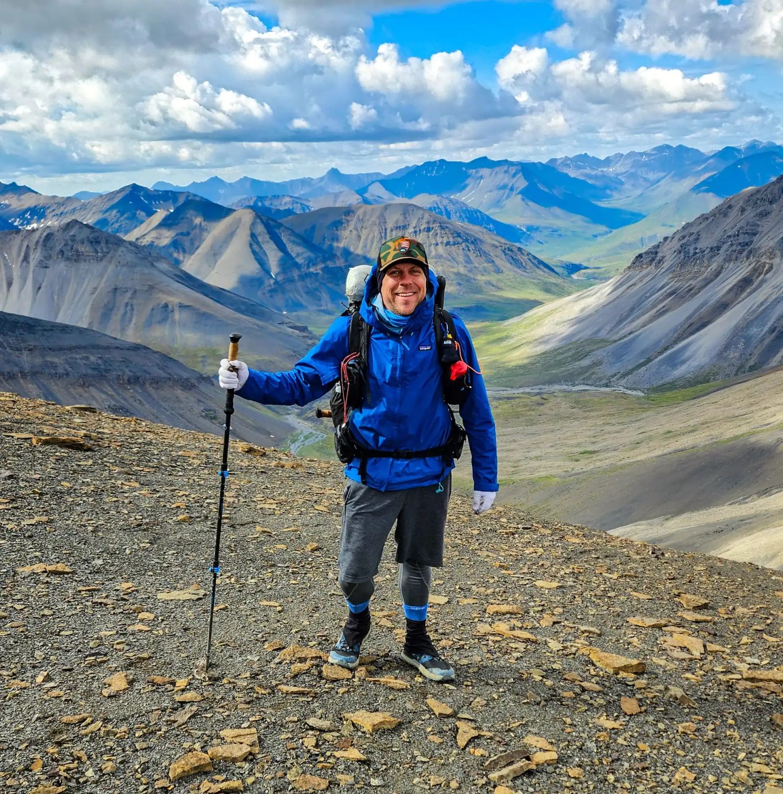 Eric Kufrin stands with hiking poles and backpack on rocky terrain overlooking vast mountain valleys and peaks in Gates of the Arctic National Park