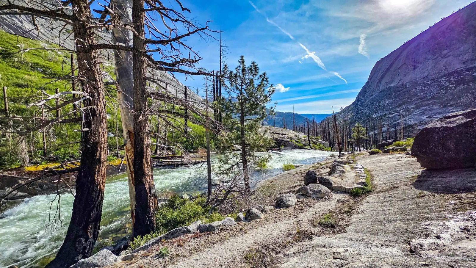 Rocky granite landscape with rushing river, burned trees, and mountain cliffs under blue sky with wispy clouds
