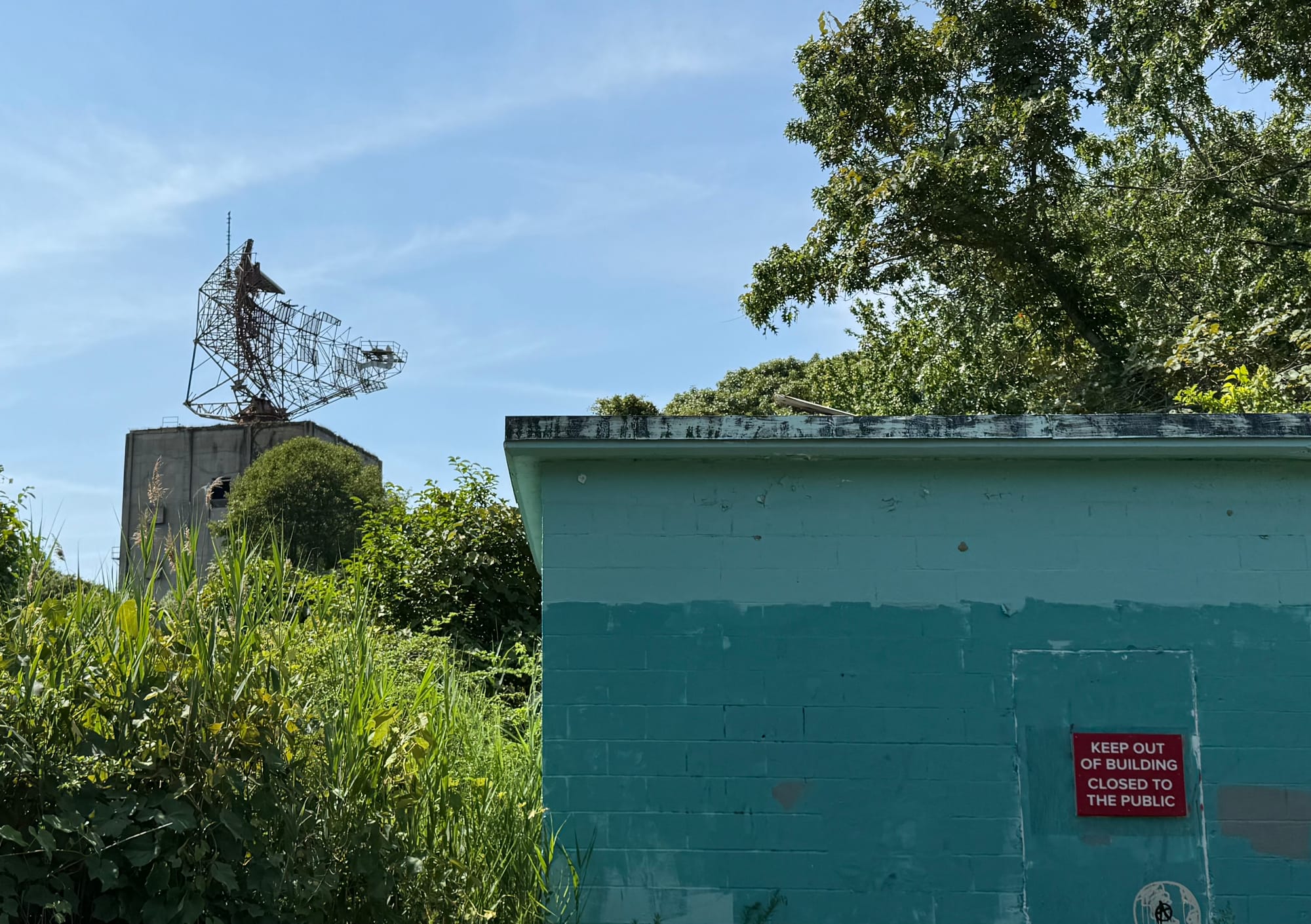 A photograph of a radar station against a bright blue sky with high cumulus clouds. Weeds and vines overgrown a small concrete building in the foreground. A Sign tacked to the door says KEEP OUT OF BUILDING CLOSED TO PUBLIC In all capital letters. The paint is fading from the building and the gigantic radar dish panels have all fallen away revealing a skeleton reminiscent of fishbones. 