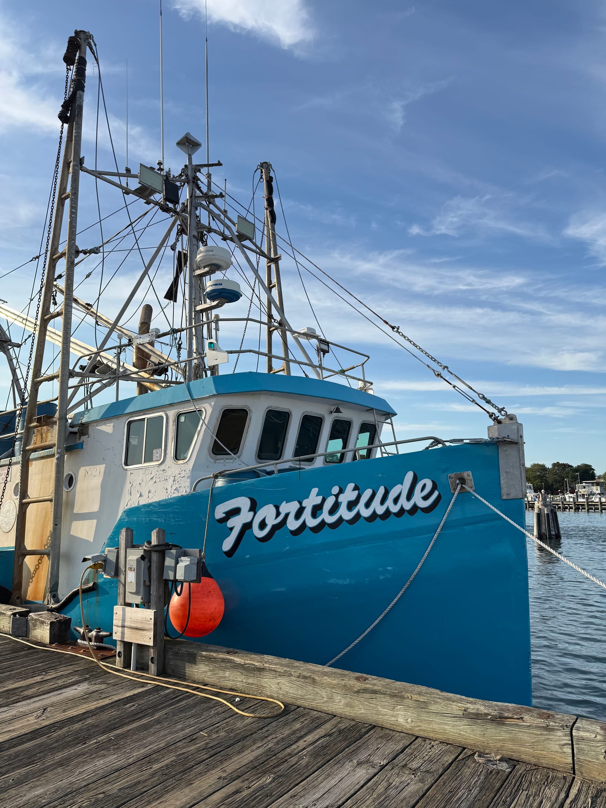 A photograph of a charming fishing vessel called Fortitude, in Greenport, Long Island, NY. It has a cheerful prow. The script proclaiming its name is optimistic and neatly painted. While the paint is chipped and there is rust, it looks clean and scrubbed and otherwise well maintained. 