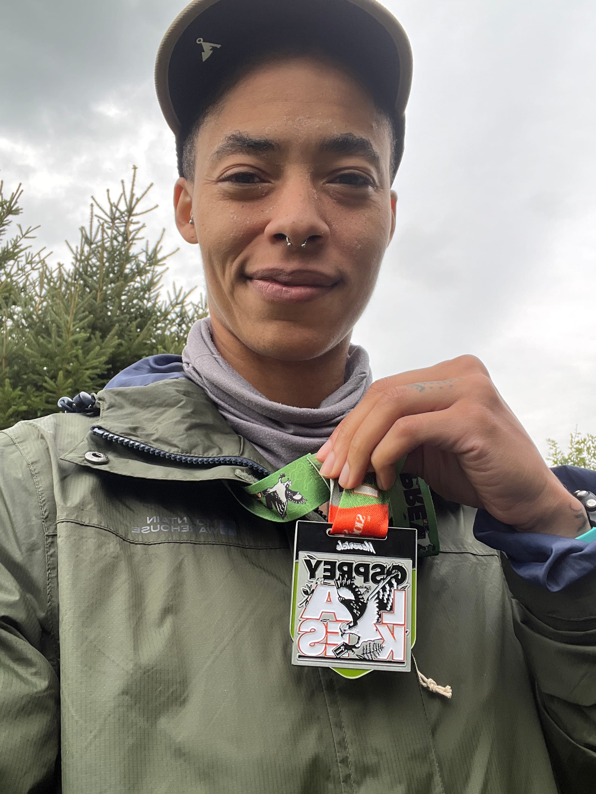 c. l. clark in a rain jacket with salt crusting their face, holding a race medal that they are wearing around their neck.