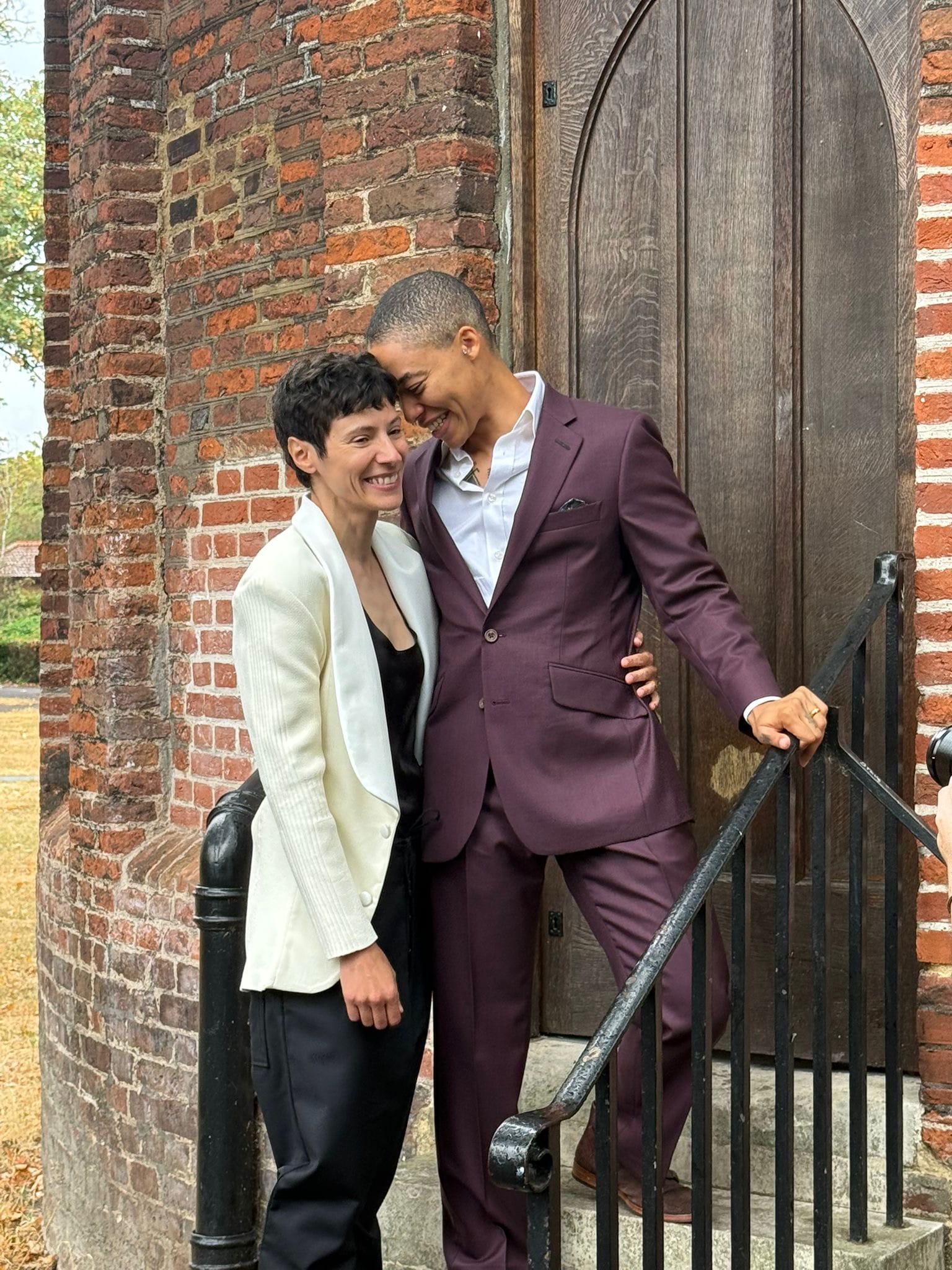 two masc queers making heart eyes at each other, one in a cream and black suit, the other in a toasted plum suit