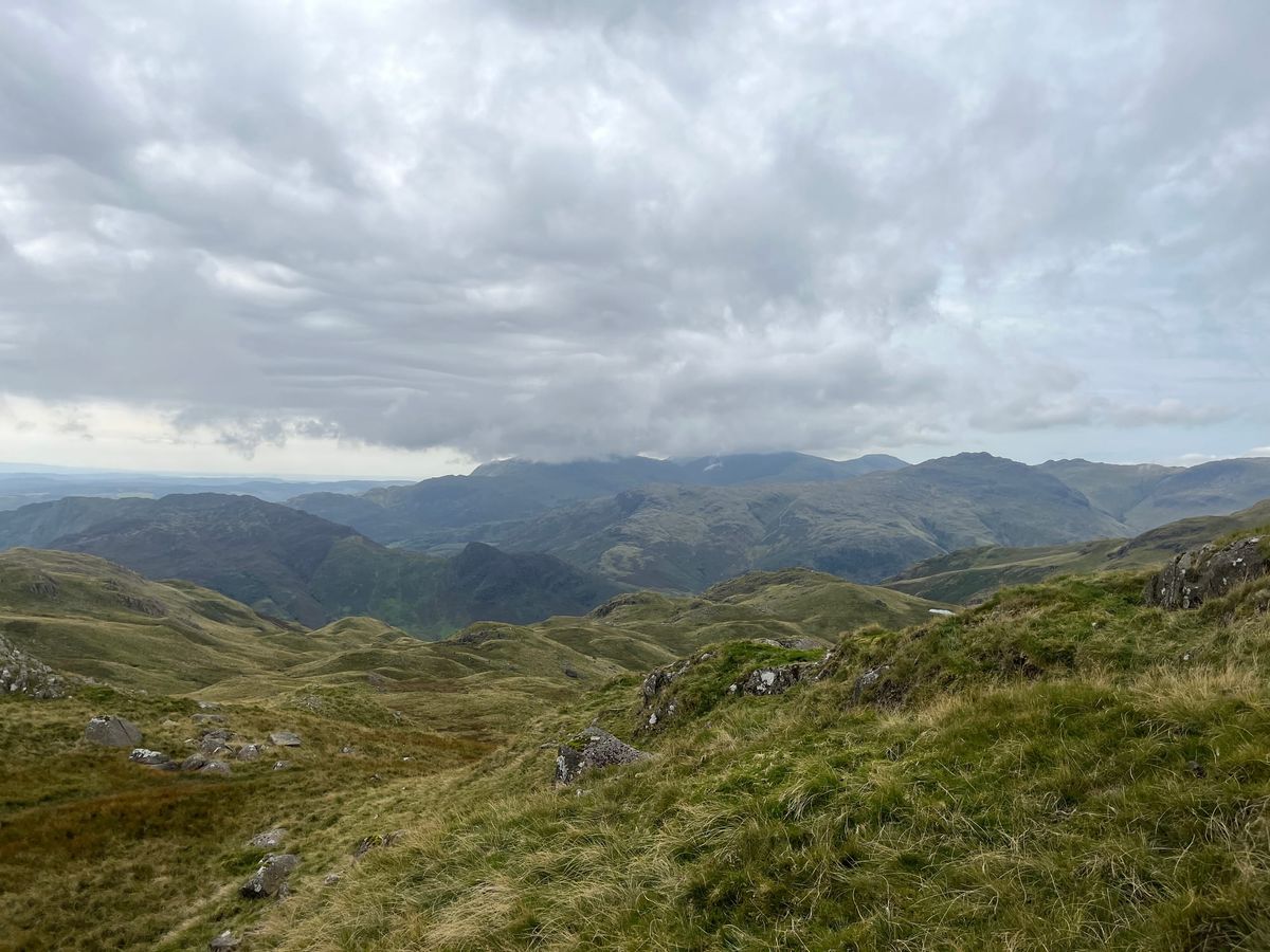 a view of a very cloudy sky and grassy fells from the top of another fell.