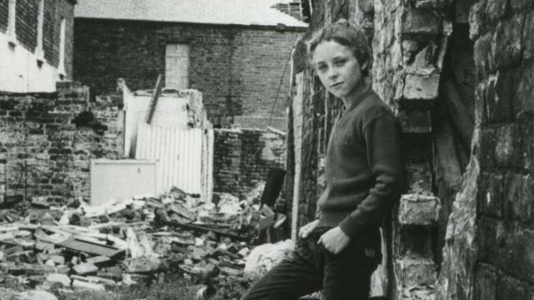 A black and white photo of a young Irish person stands amid rubble next to a bombed building.