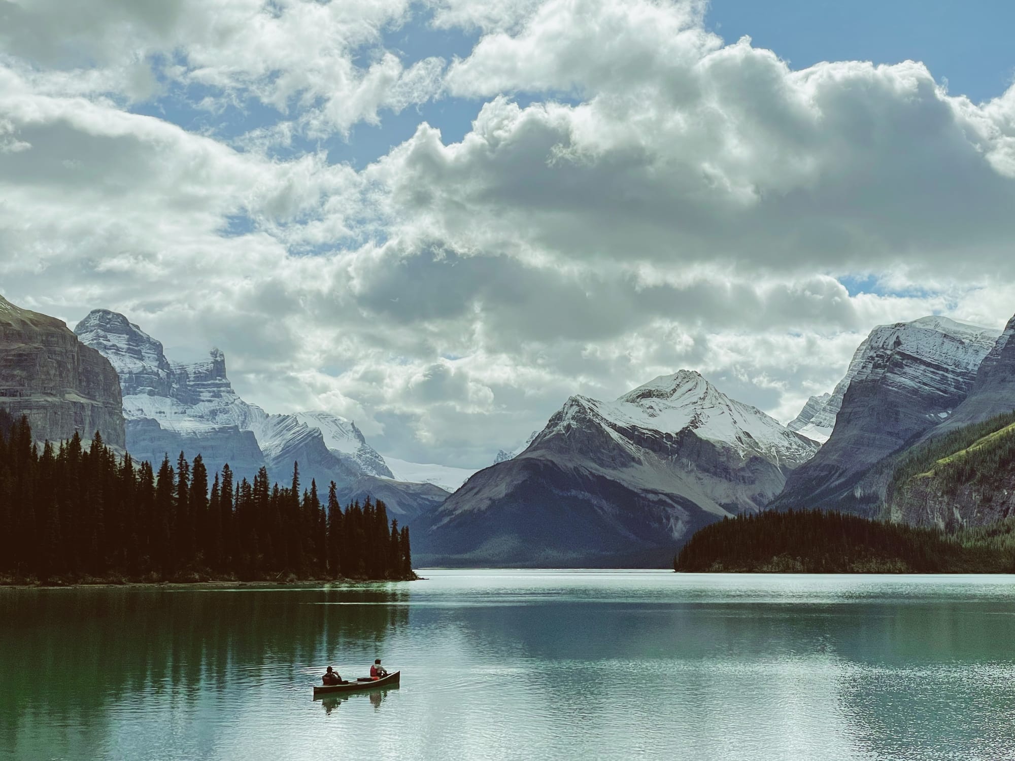 In the foreground, two people sitting on a canoe on a vast lake look out at a panorama of towering rocky mountains dusted with snow under a partially-cloudy sky.
