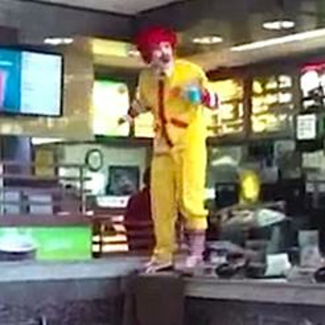 A pretty deep-fried JPEG of an individual in a bright yellow Ronald McDonald costume standing on the counter of a McDonald's.