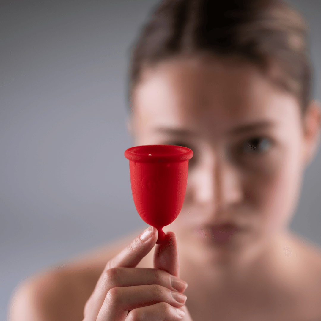 Detailní záběr na červený silikonový menstruační kalíšek, který drží žena v prstech před sebou, s rozostřenou tváří v pozadí.