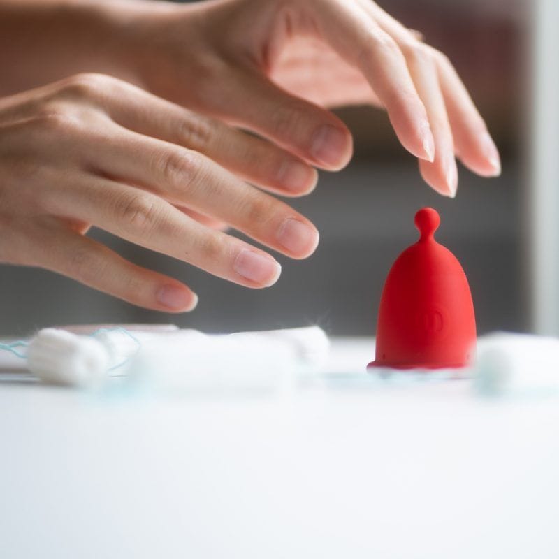 Detailní záběr na ruce nad stolem, kde stojí červený menstruační kalíšek Whoop·de·doo obklopený rozostřenými jednorázovými tampony.